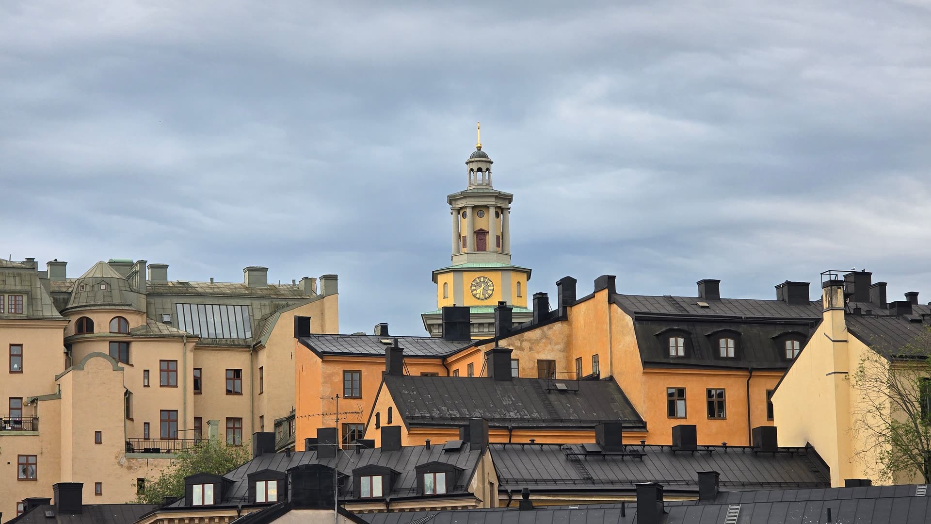 Söders tak sedda i nivå med varandra: svart plåt, rader av skorstenar, takkupor och stegar, med ett gult klocktorn över.
